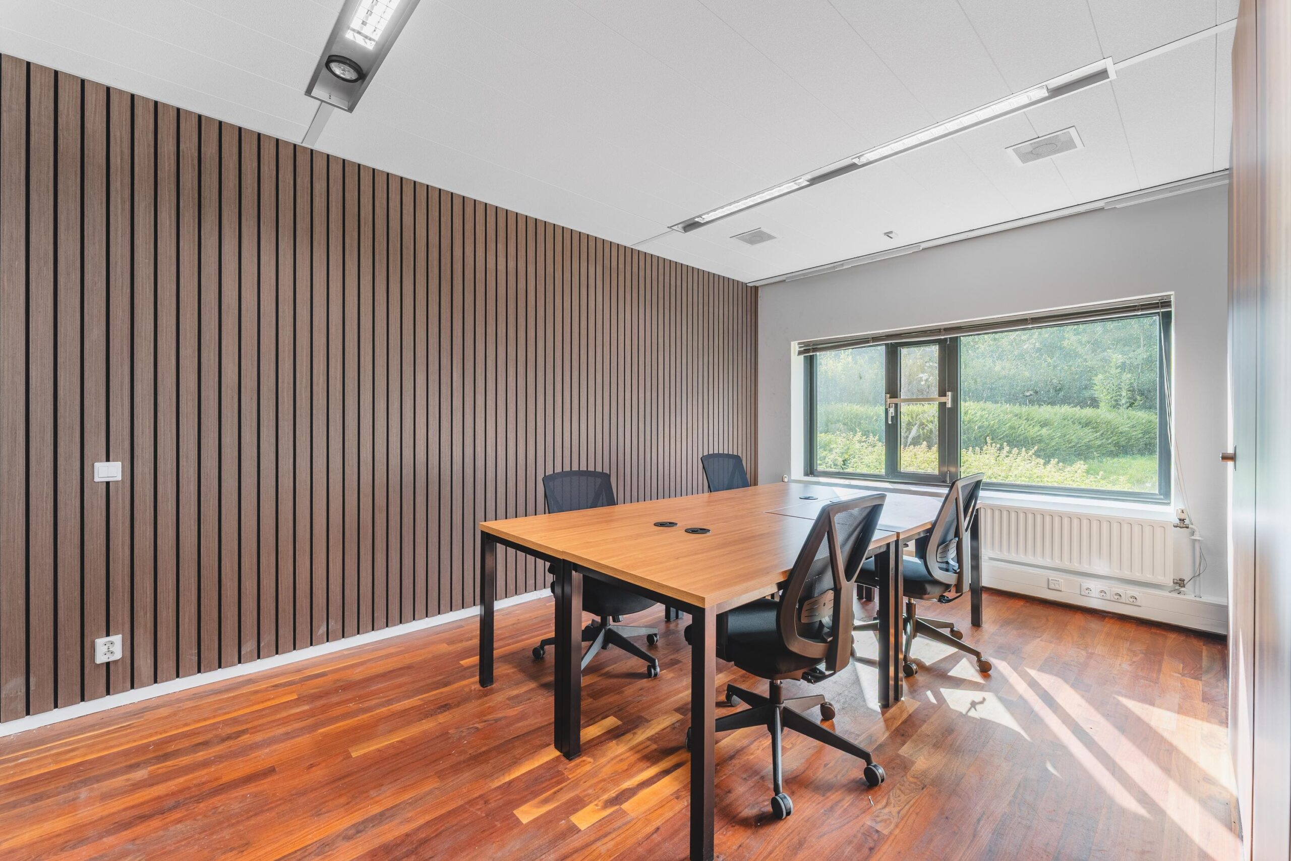 Vergaderruimte met houten vloer, moderne tafel, vier bureaustoelen en een groot raam met uitzicht op groen aan de Prof. W.H. Keesomlaan.